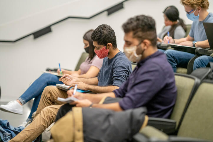 College students sitting in a lecture hall on campus, all are wearing masks to protect from the transfer of germs.